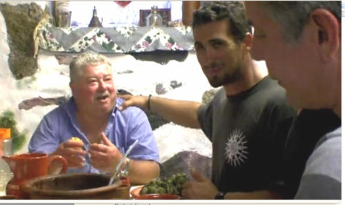 Anthony Bourdain on the right and a guy wearing a 'Fabric of Life' shirt next to him, somewhere in Greece
